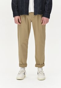 Jeansjacke über einem weißen Hemd, kombiniert mit khakifarbenen Baumwollhosen, die an den Bündchen hochgekrempelt sind. Sneaker mit weißem Grund und navyblauen Akzenten vervollständigen das Outfit.