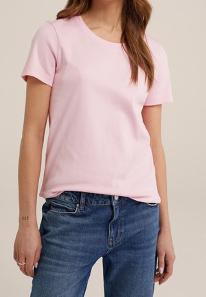 Kvinde iført en ensfarvet lyserød T-shirt med korte ærmer, let stoppet ned i højtaljede blå denimjeans, stående foran en neutral baggrund.