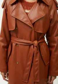 Bruine faux leren trenchcoat met een ceintuur in de taille, brede revers, dubbele knopen en zijzakken. Gladde textuur en nauwkeurige pasvorm.