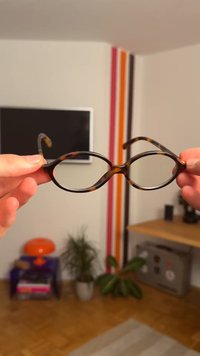 Okuliare s oválnym rámom z plastu so vzorom korytnačiny. Tenké ramená sa rozširujú von. Priehľadné šošovky. Ruky držia okuliare proti neutrálnemu pozadiu.