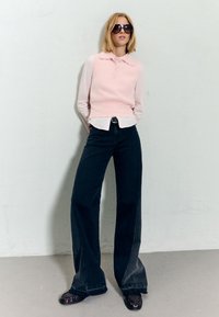 Žena nosi velike crne sunčane naočale, ružičasti bezrukavnik preko bijele košulje, široke crne traperice, stoji naslonjena na običnu zid.