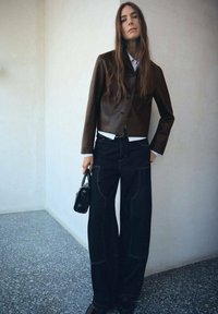 Chaqueta de cuero marrón corta, camisa blanca, jeans oscuros de gran tamaño con costuras en contraste y un pequeño bolso negro. Textura suave y ajuste entallado.