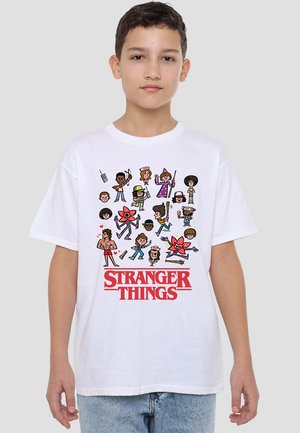 Junge trägt ein weißes T-Shirt mit Cartoon-Charakteren aus Stranger Things und rotem Logo und steht vor einem hellgrauen, einfarbigen Hintergrund.