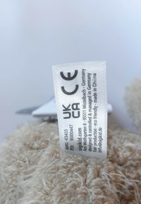 Clothing tag on beige textured fabric showing CE, UKCA marks and text about design in Germany and manufacturing in China.
