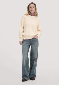 Cremfarbener Rippstrickpullover mit langen Ärmeln, über einem rot-weiß gestreiften Rollkragenpullover getragen; hellblaue weite Jeans, dunkelbraune Schuhe.