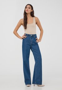 Body suit color marrone chiaro con scollatura quadrata abbinato a jeans di denim blu scuro a vita alta e gamba larga. Texture lisce e dettagli di cucitura sottili.