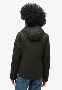 Dunkelgrüne Kapuzenjacke mit strukturiertem Material, elastischen Bündchen und geradem Saum, getragen über hellblauen Jeans.