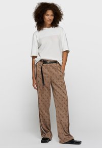Pantalones de pierna ancha con estampado marrón, cinturón negro y detalles de cremalleras plateadas, combinados con una camiseta blanca holgada. La modelo está de pie frente a un fondo liso.