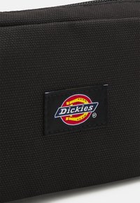 Bolsita de tela negra con una superficie texturizada y una etiqueta rectangular visible con el logo de "Dickies" en rojo, amarillo y azul.