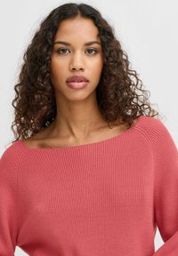 Pulover z dolgimi rokavi, rebrasto pleten v roza odtenku z širokim ovratnikom in nekoliko skrajšanim krojem, narejen iz mehkega, teksturiranega materiala.