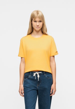 Ung kvinde med langt blondt hår iført en ensfarvet gul t-shirt og blå jeans med en hvid snor, stående foran en hvid baggrund.