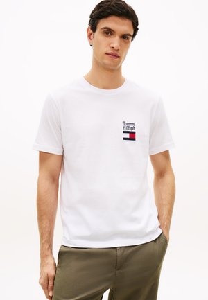 Noor mees, kes kannab valget Tommy Hilfigeri t-särki rinnal logoga, kombineeritud oliiviroheliste pükstega, seisab ühevärvilise valge tausta ees.