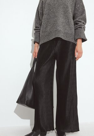 Sorte plisserede bredbenede bukser lavet af skinnende stof, parret med en løs grå strikket sweater og en sort tote taske. Minimalistisk design.