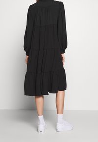 Robe noire à volants avec des manches longues, en tissu texturé et à coupe décontractée. Portée avec des baskets blanches et des chaussettes de cheville.