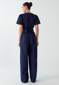 Modra overal z kratkimi napihnjenimi rokavi, zavezanim pasom in širokimi hlačami, z gladko teksturo in zadrgo na hrbtni strani.