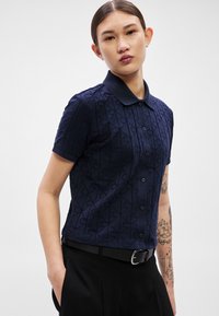 Tumši zils polo krekls ar īsām piedurknēm, teksturētu ģeometrisku rakstu, pogu aizdari un punktētu apkakli, kombinēts ar melnām biksēm un jostu.