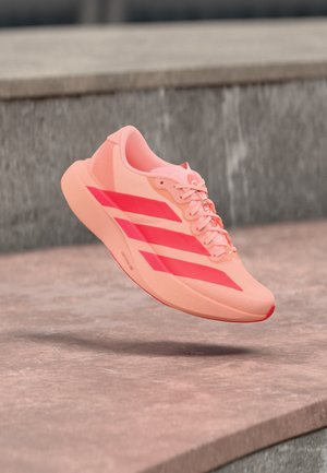 Chaussure de sport corail clair avec des rayures rouges vives et des lacets flottant au-dessus de marches en pierre dans un cadre extérieur.