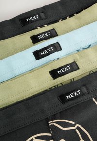 Pile de cinq articles en tissu pliés en noir, vert et bleu clair, chacun avec une étiquette noire portant la mention « NEXT » cousue près du bord.