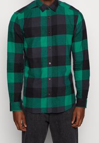 Hombre vestido con camisa de botones a cuadros verdes y negros de manga larga y vaqueros oscuros, posando contra un fondo liso.