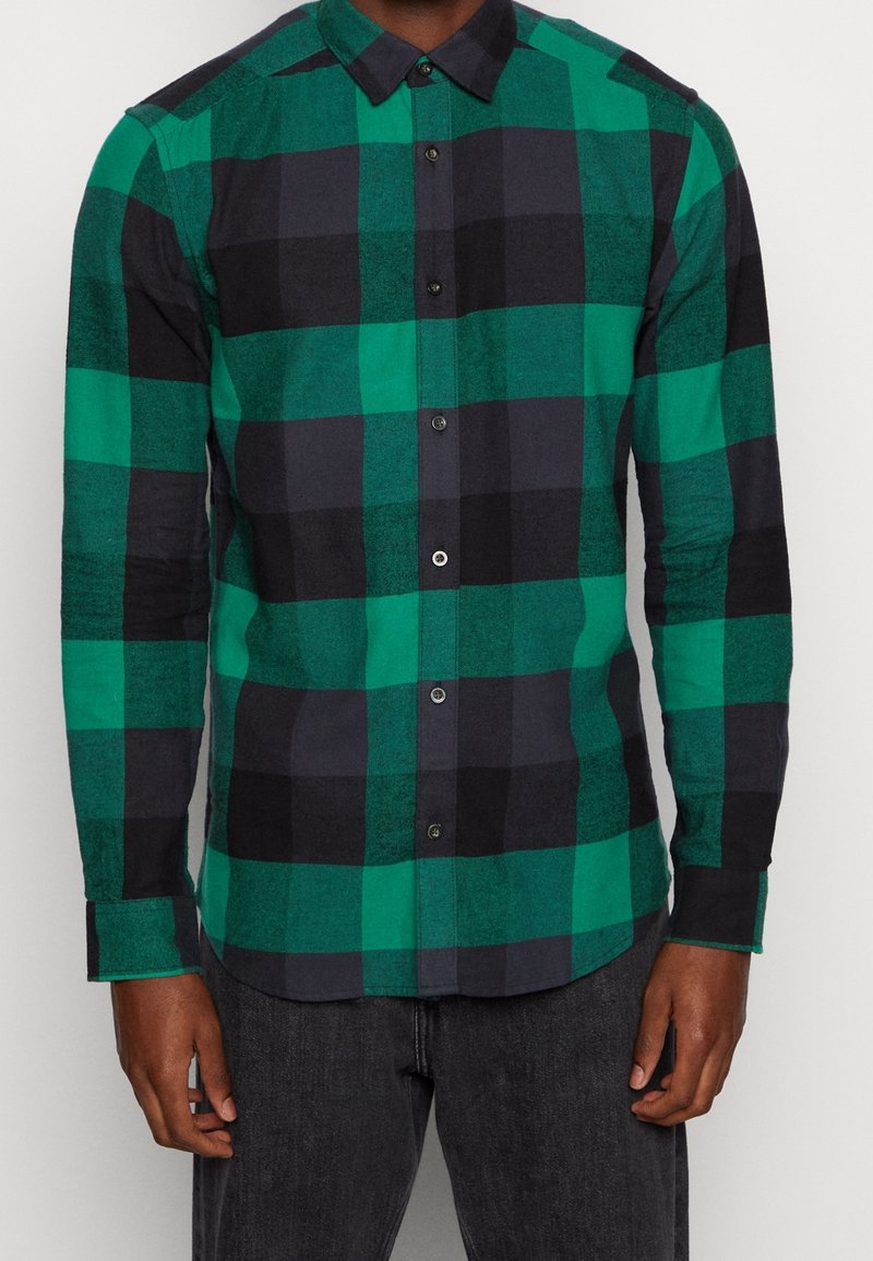 Hombre vestido con camisa de botones a cuadros verdes y negros de manga larga y vaqueros oscuros, posando contra un fondo liso.