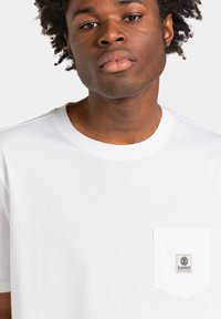 Hvid bomulds t-shirt med rund halsudskæring, korte ærmer og en lille lomme foran med et vævet mærke. Glat tekstur.