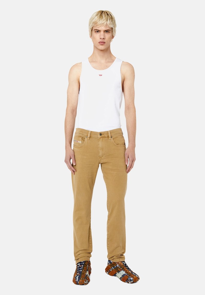 Jean beige slim, associé à un débardeur blanc. Le modèle porte des chaussures brunes à motif. L'arrière-plan est blanc uni.