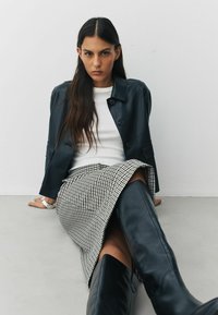 Svart läderjacka med knappar, vit ribbad tröja, hounds-tooth kjol och knähöga svarta stövlar. Modell sittande mot en neutral bakgrund.