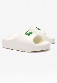 Chanclas blancas de material suave, con puntas abiertas, un logo de cocodrilo verde en la parte superior y una suela texturizada para mayor agarre.