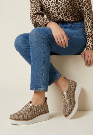 Persoon draagt een blauwe spijkerbroek, een luipaardprint shirt en beige luipaardprint slip-on platformschoenen, zittend op een lichtgekleurde bank.