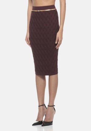 Donna che indossa una gonna aderente texturizzata di colore viola scuro, lunga fino al ginocchio, con una sottile cintura a catena dorata e sandali neri con cinturino alla caviglia e punta affusolata.