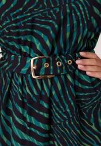 Sukienka w zielono-ciemnoniebieskie paski z pasem w talii. Pasek posiada złotą klamrę i oczka, podkreślając gładką teksturę materiału.