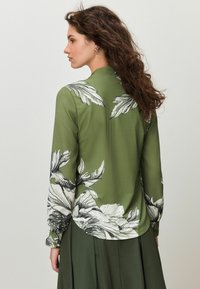 Žalia palaidinė su ilgomis rankovėmis ir apvalia apačia, puošta dideliais juodai baltais gėlių raštais, pagaminta iš minkštos, lygaus audinio.