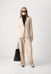 Beige tailored suit with a fitted blazer and wide-leg pants, paired with a black turtleneck and oversized black tote bag. Dark sunglasses.