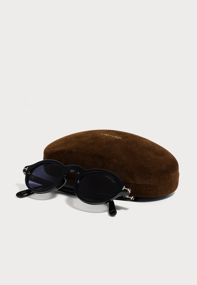 Lunettes de soleil rondes noires Tom Ford posées sur un étui en suède marron marqué Tom Ford, sur fond blanc.