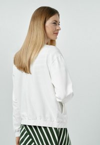 Biała, oversize'owa bluza z okrągłym dekoltem, prążkowanym ścięciem u dołu oraz bocznymi kieszeniami; noszona z zielonymi spodniami w paski. Gładka tekstura materiału.