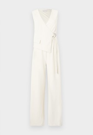 Άσπρη ολόσωμη φόρμα χωρίς μανίκια με φαρδιά πόδια, μπροστινό μέρος τύπου κρουαζέ που στερεώνεται με ασημένια αγκράφα σε σχήμα δακτυλίου και μικρή τσέπη στο πάνω αριστερό μέρος.