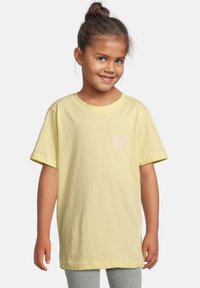 Jeune fille avec des cheveux bruns en chignon, portant un T-shirt ample jaune pâle avec un petit papillon rose sur la poitrine et un legging gris.