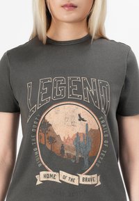 Suurendatud vaade hallile t-särgile, millel on vintage Texase kõrbe kujundus, mis sisaldab kaktusi, ratsanikku, lendavat lindu ning teksti "Legend" ja "Home of the Brave."