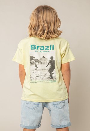 Enfant aux cheveux blonds mi-longs portant un t-shirt jaune avec un imprimé de beach soccer en noir et blanc et un short en denim clair.