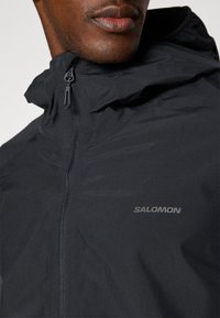 Leichte schwarze Jacke mit Reißverschluss vorne, hohem Kragen und dem "Salomon"-Logo auf der Brust. Hergestellt aus einem glatten, wasserabweisenden Material.