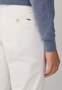 Pantalones blancos de algodón con un bolsillo trasero que cuenta con un botón beige y una etiqueta azul marino. Combinados con un suéter azul de punto.