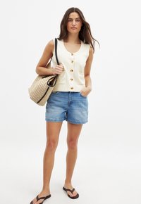 Krämbelädd utan ärmar med stora guldknappar, i kombination med blå denimshorts och bruna flip-flops. Flätad halmhandväska med svarta remmar.