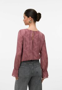 Blusa a maniche lunghe in rosa polveroso con motivo floreale testurizzato. Presenta una scollatura a chiave sul retro e maniche a campana, abbinata a jeans grigi a vita alta.