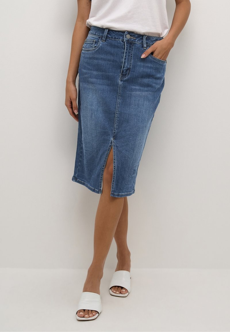 Femme portant une jupe en denim bleu mi-longue avec une fente sur le devant, un haut blanc et des sandales blanches ouvertes aux orteils, debout devant un fond uni.