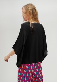 Černý lehký top s volným batwing střihem, širokým výstřihem a krátkými rukávy, doplněný barevnými vzorovanými kalhotami.