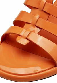 Orange patent leather sandal featuring wide, crisscross straps and a smooth, glossy finish. Rounded toe and flat sole design.