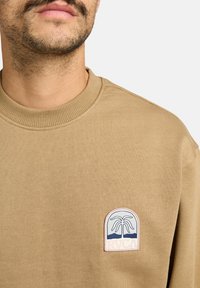 Sweatshirt marron avec col rond côtelé, arborant un patch circulaire du logo RVCA avec un palmier et des montagnes en bleu et rose.