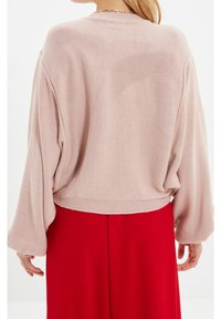 Haut tricoté rose clair avec manches longues et larges, encolure arrondie, et ourlet court, associé à une jupe rouge. Détail du tissu texturé visible.