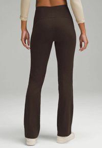 Femme portant un pantalon évasé taille haute marron foncé et des baskets blanches, debout les mains détendues le long du corps.