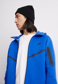 Casaco azul com fecho, detalhes em preto, bolso com fecho na manga e capuz. Estilizado sobre uma camisa branca. Complementado por um gorro de malha preto.